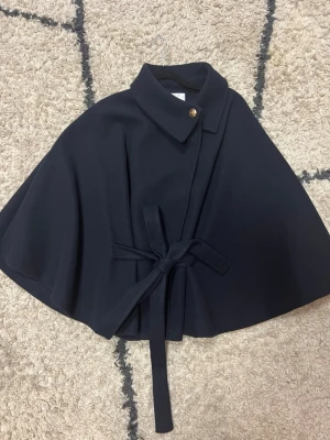 Svart cape-kappa med knytband - Stilren svart cape-kappa med klassisk krage, dekorativ knapp och knytband i midjan. Kappan har en elegant, vid siluett och är tillverkad i ett tjockare tyg som ger en lyxig känsla. Perfekt för dig som vill ha en trendig och unik ytterjacka. ( knappen längst upp har gått av men går att sy tillbaka jätte lätt) passar som både S/M