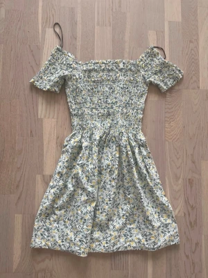 Blommig offshoulder sommarklänning - Söt kortklänning med blommigt mönster i vitt, gult och grönt. Offshoulder-modell med korta ärmar och smockad överdel som ger en bekväm passform. Kjolen är lätt utställd och klänningen är perfekt för varma dagar.