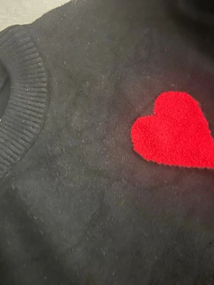 Svart stickad tröja med rött hjärta - Säljer en svart stickad tröja med ett stort rött hjärta och bokstaven A broderat på bröstet. Tröjan har ribbstickade muddar vid ärmslut och halsringning. Perfekt för dig som gillar stilrena plagg med en twist.