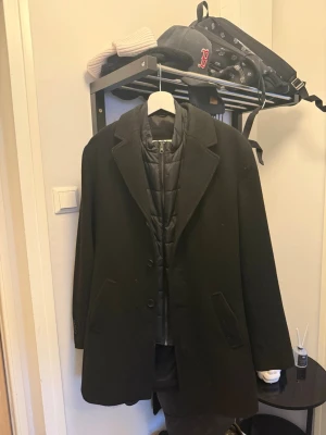 Svart kappa från bruun & stengade  - Stilren svart rock från bruun & stengade med klassisk krage och två fickor framtill. Kappan har en avtagbar quiltad innerdel med dragkedja för extra värme. Perfekt för kyliga dagar och enkel att matcha med olika outfits.