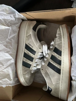 Adidas Campus beige sneakers - Säljer ett par Adidas Campus sneakers i beige mocka med svarta klassiska stripes på sidorna och vit sula. Skorna har vita skosnören och gummisula i brunt. Perfekta för dig som gillar retrostil och streetwear.
