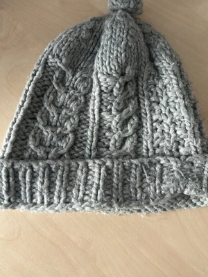 Grå stickad mössa med flätmönster - Säljer en chunky grå stickad mössa med snyggt flätmönster och uppvikt kant. Mössan har en liten tofs på toppen och är gjord i tjockt garn som känns riktigt mjukt och varmt. Perfekt för kalla dagar och ger en cool, avslappnad vibe.