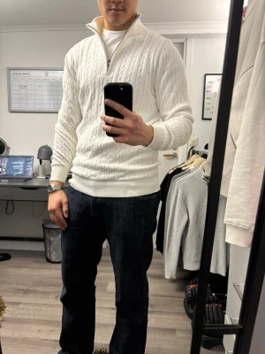 Vit kabelstickad tröja med halvzip - Snygg vit kabelstickad tröja med halv dragkedja och hög krage. Tröjan har ribbade muddar och nederkant, samt ett klassiskt mönster som ger en stilren look. Perfekt för lager på lager och passar till jeans eller chinos.