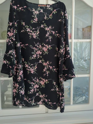 Svart blommig klänning med volangärm - Svart kortklänning med rosa och grönt blommönster. Klänningen har v-ringning och långa volangärmar som ger en romantisk vibe. Tillverkad i ett lätt och luftigt material, perfekt för varmare dagar. Storlek 16=xl