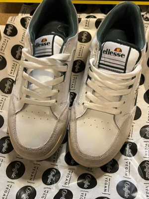 Vita Ellesse sneakers med mockadetaljer - Snygga vita sneakers från Ellesse med detaljer i mocka och grönt. Klassisk låg modell med Ellesse-logga på sidan och baksidan. ANVÄNDES cirka 3 gånger.💚