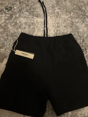 Svarta shorts från Essentials - Svarta shorts från Essentials Fear of God med diskret logga på benet och dragsko i midjan. Klassisk passform och elastisk midja för extra komfort. Perfekta för chill dagar eller streetwear-stil.