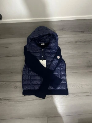 Mörkblå dunjacka från Moncler - Snygg mörkblå cardigan från Moncler med quiltat mönster, huva och dragkedja framtill. Jackan har stickade ärmar och muddar samt Moncler-logga på ena ärmen. Perfekt för kalla vinterdagar och riktigt trendig look. Storlek M-L men rekommenderar L 