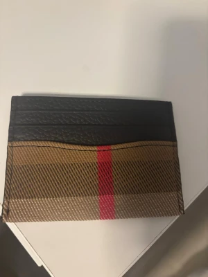 Burberry kortfodral i läder - Säljer ett stilrent kortfodral från Burberry i svart läder med klassiskt beige rutmönster och en röd rand. Perfekt för kort och ID, smidig att ha i fickan. Lyxig känsla och ikonisk design som sticker ut.