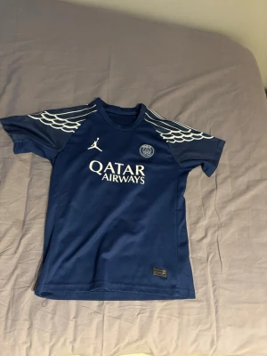 Blå PSG x Jordan matchtröja - Snygg blå matchtröja från PSG x Jordan med vita detaljer och Qatar Airways-tryck på bröstet. Tröjan har coola vita vinge-mönster på axlarna och PSG-logga på vänster sida. Tillverkad i lätt och ventilerande material, perfekt för fotboll eller träning.