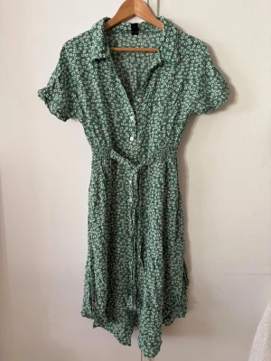 Grön blommig klänning från SHEIN - Grön kortklänning från SHEIN med vitt blommönster med grön botten, knappar framtill och knytband i midjan. Klänningen har korta ärmar, krage, kortare slitsar i sidorna och en avslappnad passform som passar perfekt till varma dagar. Använd en gång. 