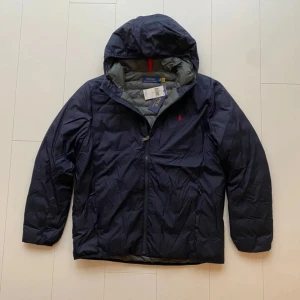 Mörkblå dunjacka från Polo Ralph Lauren - Säljer en snygg mörkblå dunjacka från Polo Ralph Lauren. Jackan är packbar och har en praktisk dragkedja. Den är perfekt för kyliga dagar och har en stilren design med det klassiska röda logotypen på bröstet, helt ny