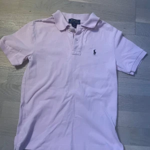 Ljusrosa pikétröja från Polo Ralph Lauren - Klassisk ljusrosa pikétröja från Polo Ralph Lauren med korta ärmar och den ikoniska broderade loggan på bröstet. Tröjan har krage och knappar vid halsen. Perfekt för en clean och stilren look. Storlek: S i barn. 