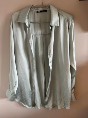 Ljusgrön satinliknande skjorta Zara - Stilren ljusgrön skjorta från Zara i ett satinliknande material. Skjortan har klassisk krage, knappar framtill och långa ärmar med manschetter. Perfekt för en trendig och avslappnad look.