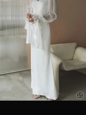 Vit långklänning med transparenta ärmar - Elegant vit långklänning med fyrkantig halsringning och transparenta långärmar. Ärmarna har dekorativa knytband vid handlederna och klänningen har en smickrande, rak siluett. Perfekt för speciella tillfällen där du vill känna dig extra fin.