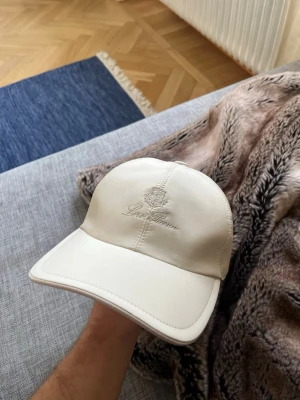 Vit keps från Loro Piana - Snygg vit keps från Loro Piana med broderad logga framtill. Kepsen har böjd skärm, panelkonstruktion och elastiskt band bak för skön passform. Tillverkad i ett slätt, exklusivt material som ger en clean och stilren look.