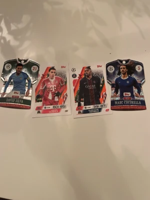 Fotbollssamlarkort Topps Champions League - Fyra fotbollssamlarkort från Topps Champions League-serien. Korten har spelare i olika lagtröjor, bland annat Chelsea, PSG, Bayern München och Real Sociedad. Två av korten är formade som tröjor och har glittriga detaljer. Perfekt för samlare av fotbollskort.
