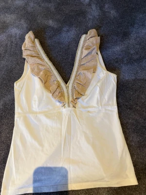 Vit topp  - Beige/guldiga volangdetaljer runt urringningen. Det finns en mindre fläck framtill (se bild ovan). Fläcken kan eventuellt gå bort med behandling, men ingen jag kan garantera - därför säljs den för ett lägre pris. Köpt utomlands! 