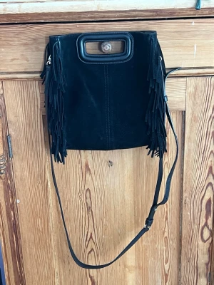 Svart axelväska med fransar från Mq - Snygg svart axelväska i mockaimitation från maje med långa fransar på sidorna och ett coolt handtag upptill. Väskan har justerbar axelrem och silverfärgade detaljer. Perfekt för dig som gillar boho-stil och vill ha en unik accessoar.