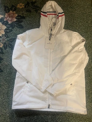 Vit vindjacka från Moncler med huva - Snygg vit vindjacka från Moncler med dragkedja, två fickor med tryckknappar och justerbar huva med röda och blå ränder. Jackan har Moncler-logga på ärmen och är tillverkad i lätt, vindtätt material. Perfekt för en stilren och sportig look.