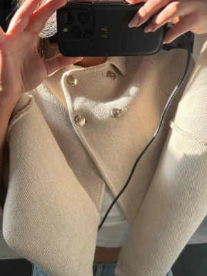 Beige poncho med guldknappar - Trendig beige poncho jacka med kort, boxig passform och vida ärmar. Jackan har dubbla rader med dekorativa guldknappar framtill och är stickad i ett mjukt material. Perfekt att slänga över en topp för en cool och avslappnad look.