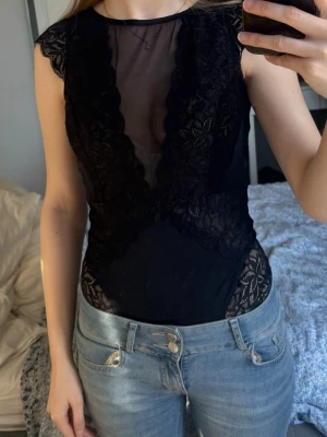 Svart spetsbody med meshdetaljer - Snygg svart body med spets och meshdetaljer. Djup urringning framtill, korta ärmar och vackra blommiga spetskanter. Står storlek 40 i men passar mig som har xs/s
