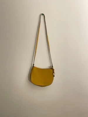 Gul axelväska i läder - Cool gul axelväska i slätt läder med enkel design. Väskan har en rundad form och en liten metallring med knutdetalj på sidan. Perfekt för dig som vill ha en färgklick till din outfit.