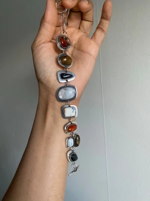 Armband med färgglada stenar - Unikt armband med stora, ovala och rektangulära stenar i olika färger som röd, brun, svart, vit och grå. Stenarna är infattade i silverfärgad metall och sitter ihop med länkar. Perfekt för dig som gillar statement-smycken och vill sticka ut.