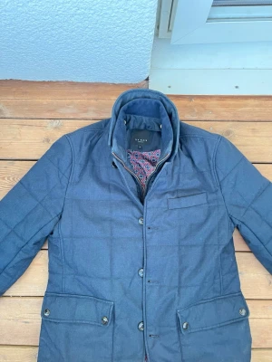 Quiltjacket Ted baker - En grå quiltjacka från Ted baker i storlek S. Jackan är i ett mycket bra skick och har en väldigt intressant interiör i ett röd/blått påfågelsmönster. Jackan har även en typ av väst på insidan med en krage som går att ta av och sätta på. Hör gärna av er om ni har några frågor☺️