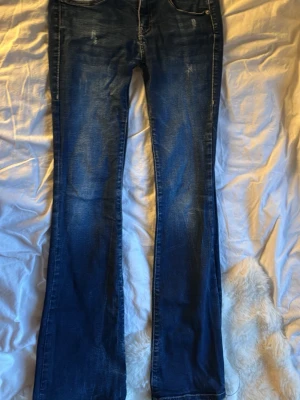 Blå bootcut jeans med slitningar - Säljer ett par lågmidjade blåa bootcut jeans med snygga slitningar och tvättade detaljer. Jeansen har klassisk femficksdesign och är tillverkade i denim med lätt stretch. Perfekta för dig som gillar en avslappnad men trendig look. Endaste skadan är den längst ner. 
