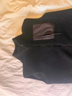 Svart fleeceväst med bröstficka - Säljer en svart fleeceväst i teddy-material med hög krage och dragkedja framtill. Västen har en snygg bröstficka i nylon med dragkedja som detalj. Perfekt att slänga över en hoodie eller tröja för extra värme och stil.