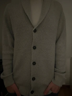 Grå kofta från Lindbergh - Mysig ljusgrå ribbstickad kofta med stora svarta knappar och bred sjalkrage. Koftan har långa ärmar och en avslappnad passform, perfekt för lager på lager. Passar till både jeans och t-shirt för en chill look.