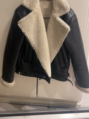 Svart teddyjacka från Gina Tricot - Snygg svart teddyjacka från Gina Tricot med stora kragar och foder i beige fuskpäls. Jackan har detaljer i läderimitation, bälte nertill och breda slag. Perfekt för kalla dagar när du vill vara både varm och trendig.