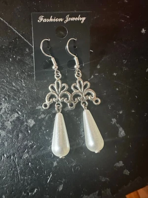 Silverfärgade örhängen med pärldroppar - Snygga örhängen i silverfärgad metall med dekorativt ornament och droppformade pärlor i vitt. Hängande modell med krokfäste, perfekt för att ge en elegant touch till din stil. Materialet är troligen metall och plast.