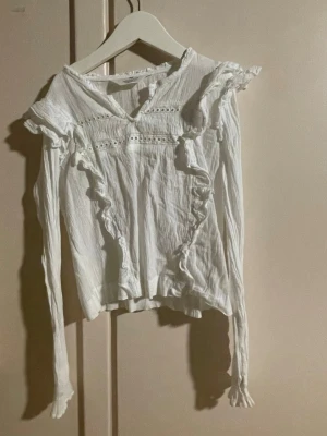 Vit volangblus med spetsdetaljer - Superfin vit blus med långa ärmar, volanger längs axlar och framsida samt spetsdetaljer vid bröstet. Tillverkad i ett lätt, krinklat tyg som ger en avslappnad men ändå söt look. Perfekt att styla med jeans eller kjol för en trendig outfit.