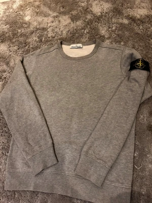 Grå sweatshirt från Stone Island - ( Öppen för BUD ocy FRÅGOR). Säljer en stilren grå sweatshirt från Stone Island med klassisk rund halsringning och ikonisk logga på vänster ärm. Tröjan har ribbade muddar vid ärmslut och nederkant. Perfekt för en avslappnad och trendig look. 