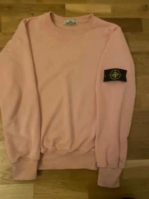 Rosa sweatshirt från Stone Island - Säljer en rosa sweatshirt från Stone Island med klassisk rund hals och ribbade muddar. Tröjan har den ikoniska svarta patchen med gul kompass på vänster ärm. Perfekt för dig som gillar streetwear och vill ha något stilrent.