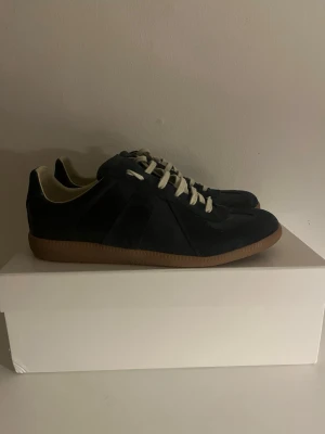 Mörkblåa sneakers från Maison Margiela - Snygga mörkblå sneakers från Maison Margiela med klassisk design och vita snören. Skorna har en ovandel i mocka och skinn, samt en brun gummisula. Perfekta för dig som gillar stilrena och tidlösa sneakers med en lyxig känsla.