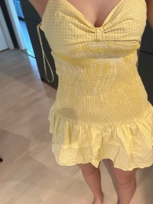 Gul kort sommarklänning med volang - Söt gul kort klänning med smala axelband och volangkant nertill. Klänningen har ett smått rutigt mönster och smockad midja som ger en bekväm passform. Perfekt för varma dagar och har en feminin, ungdomlig stil.