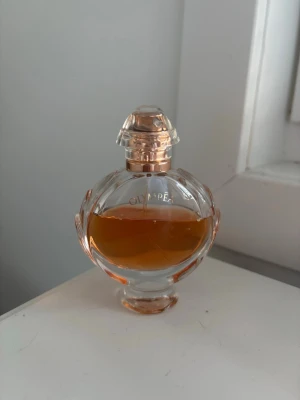 Olympea parfym från Paco Rabanne - Elegant parfymflaska från Paco Rabanne, Olympea. Flaskan är rundad med detaljer i roséguld och genomskinligt glas som visar den bärnstensfärgade vätskan. Lock i matchande roséguld och glas. Perfekt för dig som gillar lyxiga dofter. 
