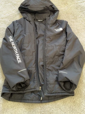 Svart dunjacka från The North Face - Snygg svart dunjacka från The North Face med huva och dragkedja. Jackan har logga på bröstet och ärmen samt reflekterande detaljer på ärmarna. Perfekt för kalla vinterdagar och har en modern, sportig look.