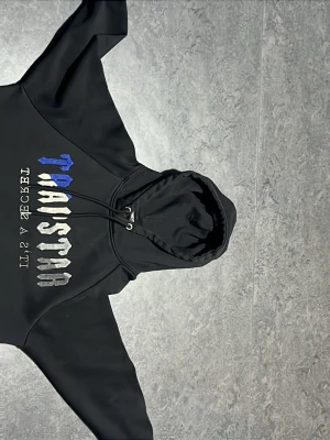 Svart Trapstar hoodie - Svart hoodie från Trapstar. Framsidan har ett iögonfallande tryck i blått, vitt och grått med texten 'Trapstar' och 'IT'S A SECRET'. Tillverkad i mjukt material, perfekt för en avslappnad streetwear-look. Hoodiens snöre är borta och den är medium fast den blev mycket smalare när jag tvättade den och därför säljer jag den.