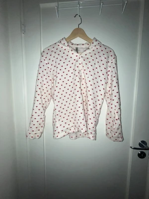 Vit pyjamaströja med röda hjärtan - Söt vit pyjamaströja med långa ärmar och små röda hjärtan över hela tyget. Tröjan har knappar framtill och är gjord i ett mjukt, strukturerat material som känns skönt mot huden. Perfekt för mysiga kvällar hemma.