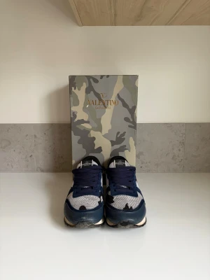 Valentino Garavani camo sneakers - Snygga sneakers från Valentino Garavani med blå, vita och svarta camo-detaljer. Ovandelen är i mesh och mocka med vita och mörkblå partier samt svart sula. Klassisk låg modell med vita och svarta detaljer och cool design.