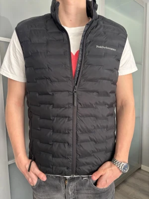 Svart dunväst från Peak Performance - Snygg svart dunväst från Peak Performance med diskret logga på bröstet. Västen har hög krage, dragkedja framtill och quiltad design. Perfekt att slänga över en hoodie eller t-shirt för en chill och sportig look.
