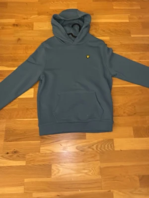 Blå hoodie från Lyle & Scott - Snygg blå hoodie från Lyle & Scott med klassisk huva och känguruficka. Diskret logga med gul fågel på bröstet. Mjuk bomullskänsla och ribbade muddar vid ärmslut och nederkant. Perfekt för chill dagar och enkel att matcha med jeans.