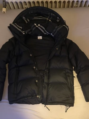 Svart pufferjacka med huva - Säljer en svart pufferjacka från Burberry med stor vadderad huva och dragkedja framtill. Insidan av huvan har ett rutigt foder i grått och vitt. Perfekt för kalla vinterdagar. Man kan även ta bort ärmarna på jackan så att det blir till en väst.