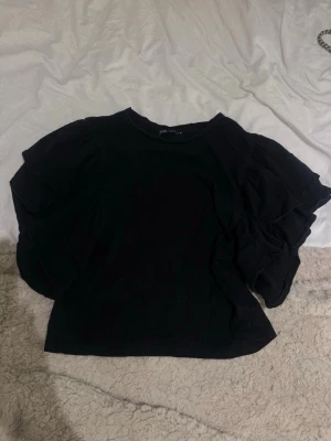 Svart volangblus med kort ärm - Svart blus från Zara med korta volangärmar och rund halsringning. Toppen har en feminin och trendig look med sina lager av volanger på ärmarna. Perfekt att styla till jeans eller kjol för en snygg outfit.