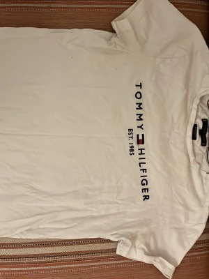 Vit Tommy Hilfiger t-shirt - Vit t-shirt från Tommy Hilfiger i slim fit-modell, tillverkad av ekologisk bomull. Framsidan har ett tryck med märkesnamnet och loggan samt texten 'Est. 1985'. Klassisk rund halsringning och korta ärmar.