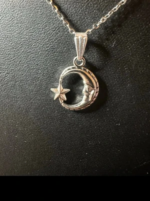 Silverhalsband med måne och stjärna - Unikt halsband i silverfärgad metall med ett hänge som föreställer en måne med ansikte och en liten stjärna. Kedjan är tunn och länkarna är diskreta, vilket gör att hänget står i fokus. Perfekt för dig som gillar mystiska och drömmiga smycken.