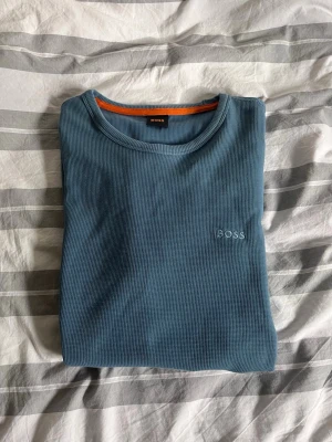 Blå långärmad tröja från BOSS - Snygg blå långärmad tröja från BOSS med diskret logga broderad på bröstet. Tröjan har rund halsringning och är gjord i ett ribbat material som ger en modern och avslappnad look. Perfekt för lager-på-lager-stil. Storleken kan vara lite för står.